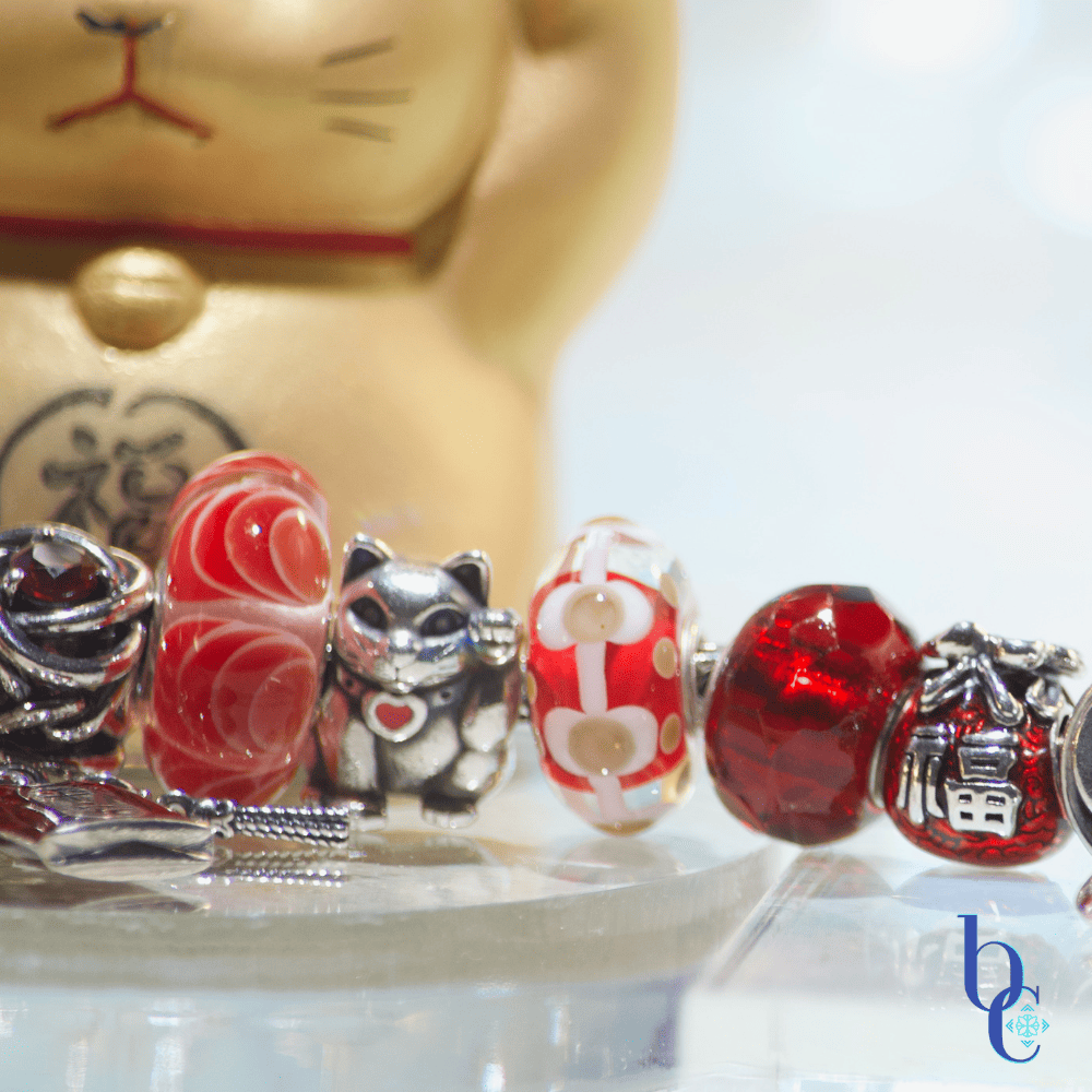 This bright red, gold and silver bracelet has charms that shout the mantra: auspiscious fortune. A Maneki neko charm is centered in the focus. Photographed on a pale background with sparkling lights and a golden metallic maneki neko figurine.
