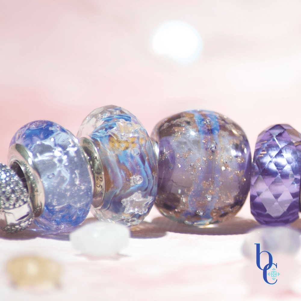 Image of an etheral ombre European bracelet design by BijouxChat.com that features am ombre of beautifully colored beads progressing from one that mimics the light of dawn to one that mimics the light of dusk.  Each glass bead is different and represents the feeling of the "time of day".  Set on a pale pink background with scattered gemstone stars around it.