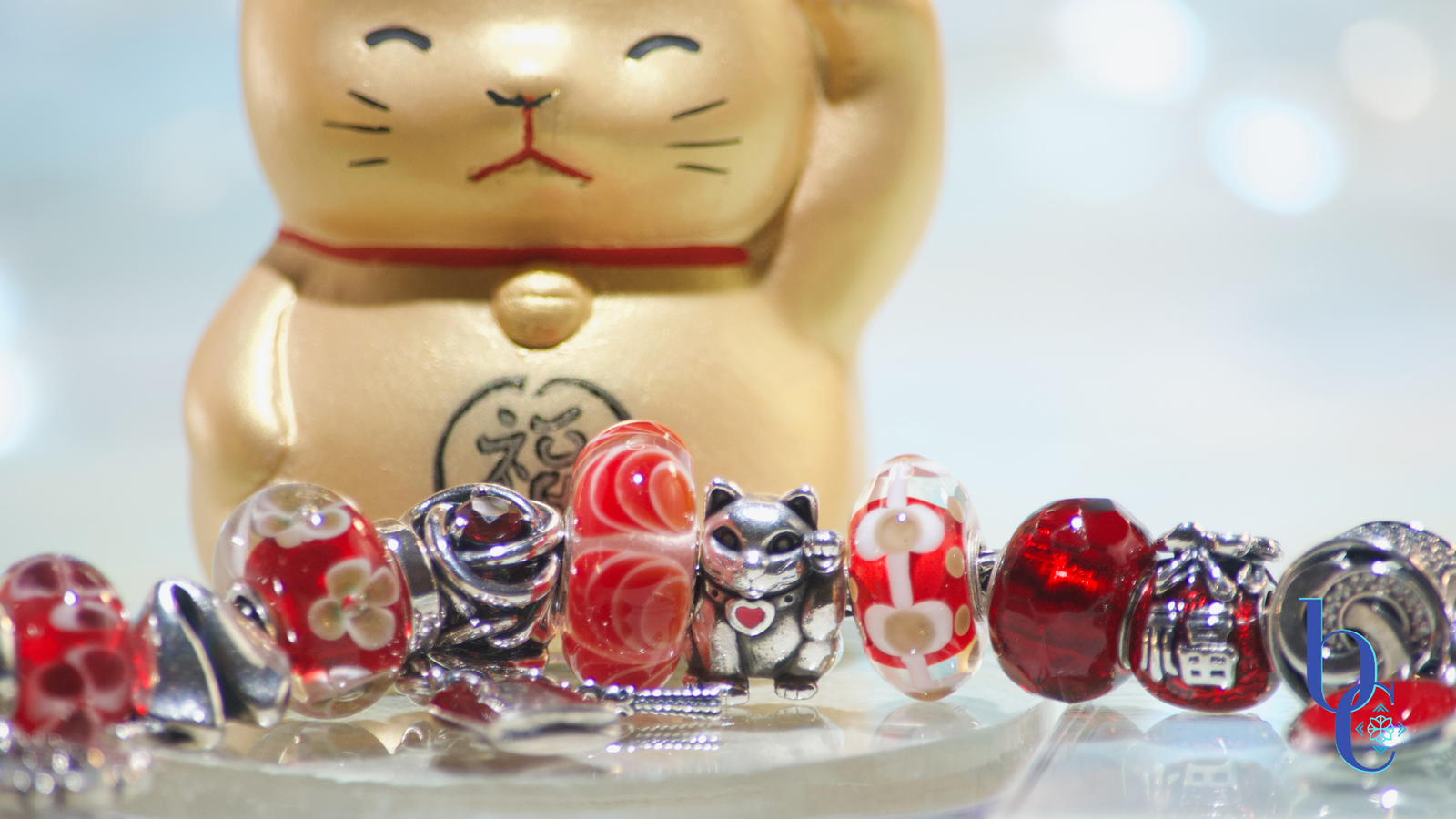 This bright red, gold and silver bracelet has charms that shout the mantra: auspiscious fortune. From a Maneki neko charm to infinity symbols in red garnets, good fortune is on your side with this design. Photographed on a pale background with sparkling lights and a golden metallic maneki neko figurine.