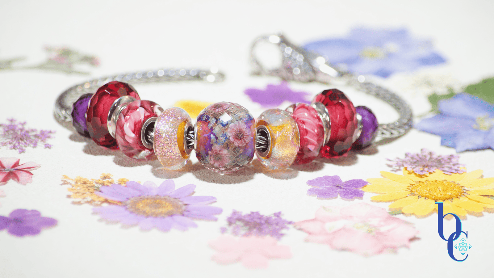 A macro shot of an European charm bracelet featuring an Elfbeads magenta Cherry Blossom Shimmering Fantasy World Fractal bead paired with orange dichroic glass charms, set against a neutral dried flower background.