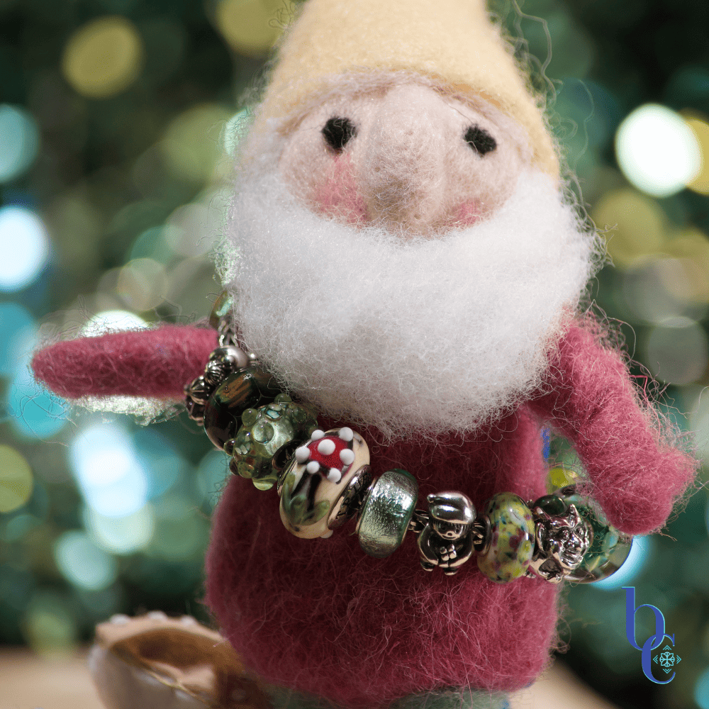 A whimsical jewelry styling scene featuring a needle-felted gnome holding a Trollbeads, Moress, Truebeadz, Black Raven Beads and Pandora forest-themed charm bracelet, set against a mossy background with glowing firefly lights.