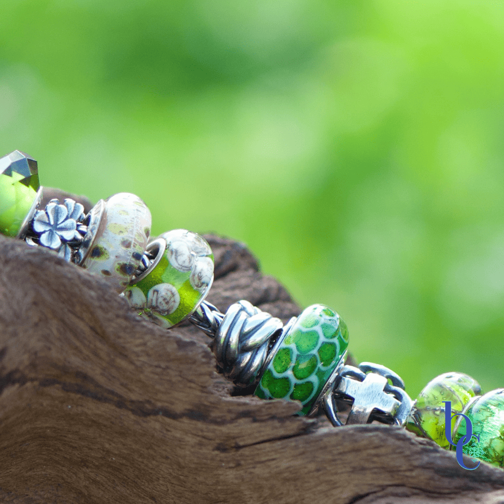 A macro featuring the Elfbeads Lucky Scales glass bead in the Bijoux Chat European bracelet design, Lucky Greens resting on rustic wood surrounded by green grass.