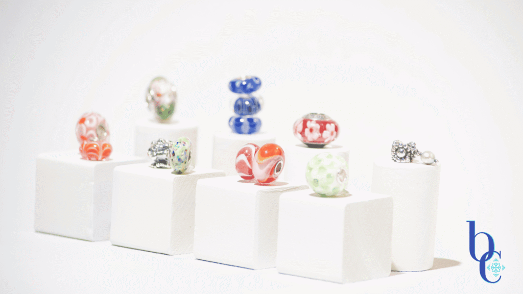 A gallery-style display of rare and OOAK charm beads arranged on clear acrylic risers against a white background, resembling a miniature museum exhibit.