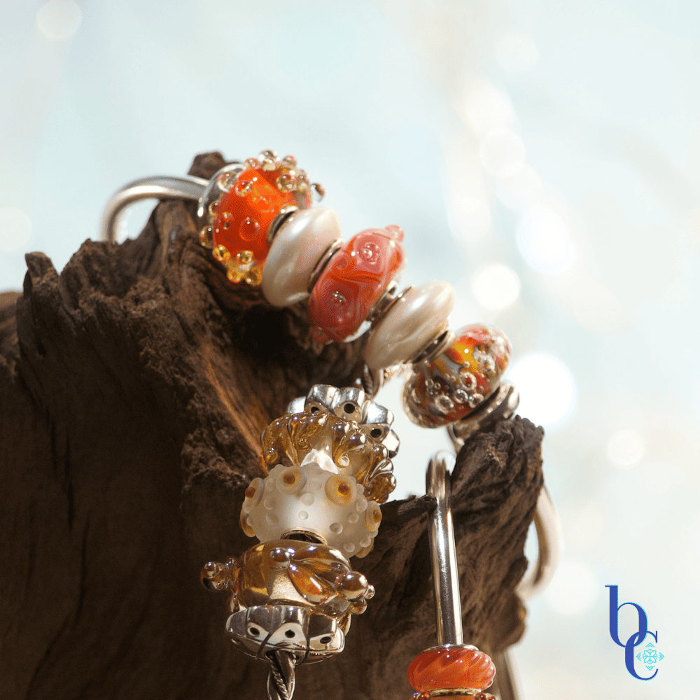 Macro photo showing a sterling silver bangles with orange and gold colored handmade glass charm beads displayed on naturally rustic dark brown wood with a pale blue background.