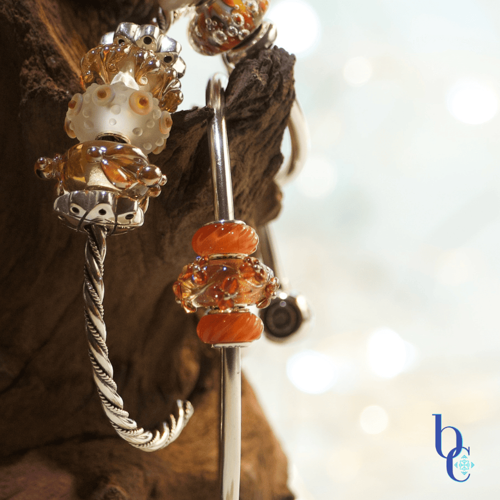 Macro photo showing a sterling silver bangles with orange and gold colored handmade glass charm beads displayed on naturally rustic dark brown wood with a pale blue background.