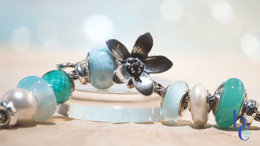 Image of A light aqua and white pearl Eurpoean charm bracelet design with glass, pearls, larimar stone, a blue agate stone and a large silver focal-the Trollbeads anemone pendant. Photographed on a natural wooden base with a pale blue background.