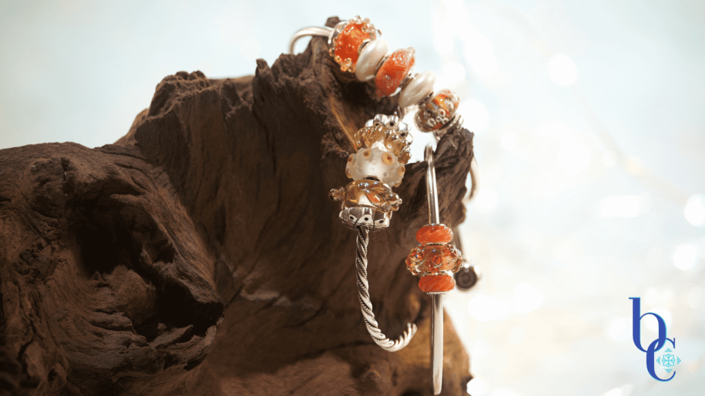 Macro photo showing a sterling silver bangles with orange and gold colored handmade glass charm beads displayed on naturally rustic dark brown wood with a pale blue background.