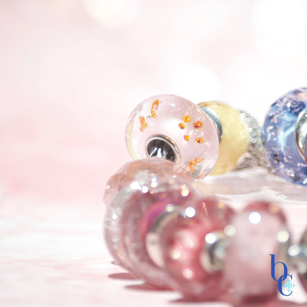 Image of an etheral ombre European bracelet design by BijouxChat.com that features am ombre of beautifully colored beads progressing from one that mimics the light of dawn to one that mimics the light of dusk.  Each glass bead is different and represents the feeling of the "time of day".  Set on a pale pink background with scattered gemstone stars around it.