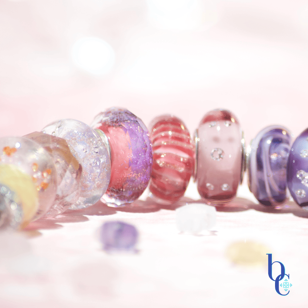 Image of an etheral ombre European bracelet design by BijouxChat.com that features am ombre of beautifully colored beads progressing from one that mimics the light of dawn to one that mimics the light of dusk.  Each glass bead is different and represents the feeling of the "time of day".  Set on a pale pink background with scattered gemstone stars around it.