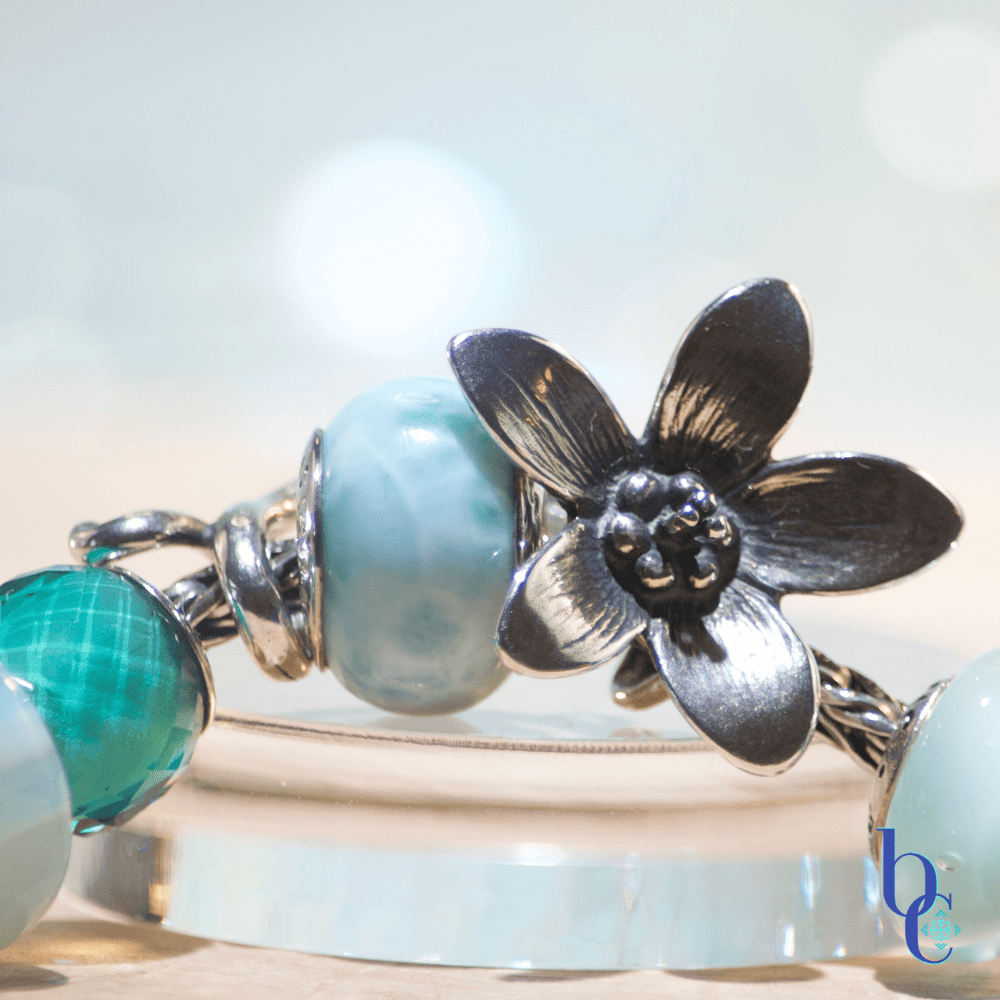 Image of A light aqua and white pearl Eurpoean charm bracelet design with glass, pearls, larimar stone, a blue agate stone and a large silver focal-the Trollbeads anemone pendant. Photographed on a natural wooden base with a pale blue background.