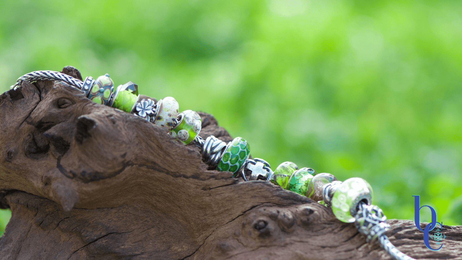 A European charm bracelet themed 'Lucky Greens' resting on rustic forest wood surrounded by fresh green grass. It features a mix of deep and light green Murano glass beads, silver Celtic knot charms, and a limited edition Trollbeads lock with lucky symbols.