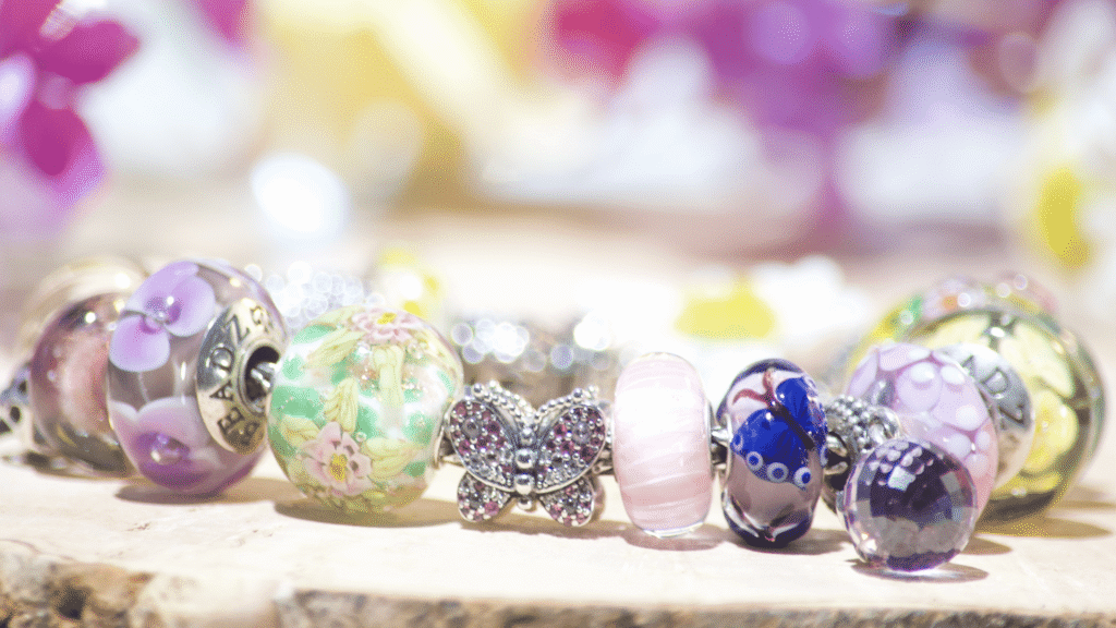 A European charm bracelet resting on whitewashed wood surrounded by fresh lilac flowers. The bracelet features chartreuse glass beads, silver butterfly charms, and pavé crystal accents, creating a 'Butterfly Garden' theme.