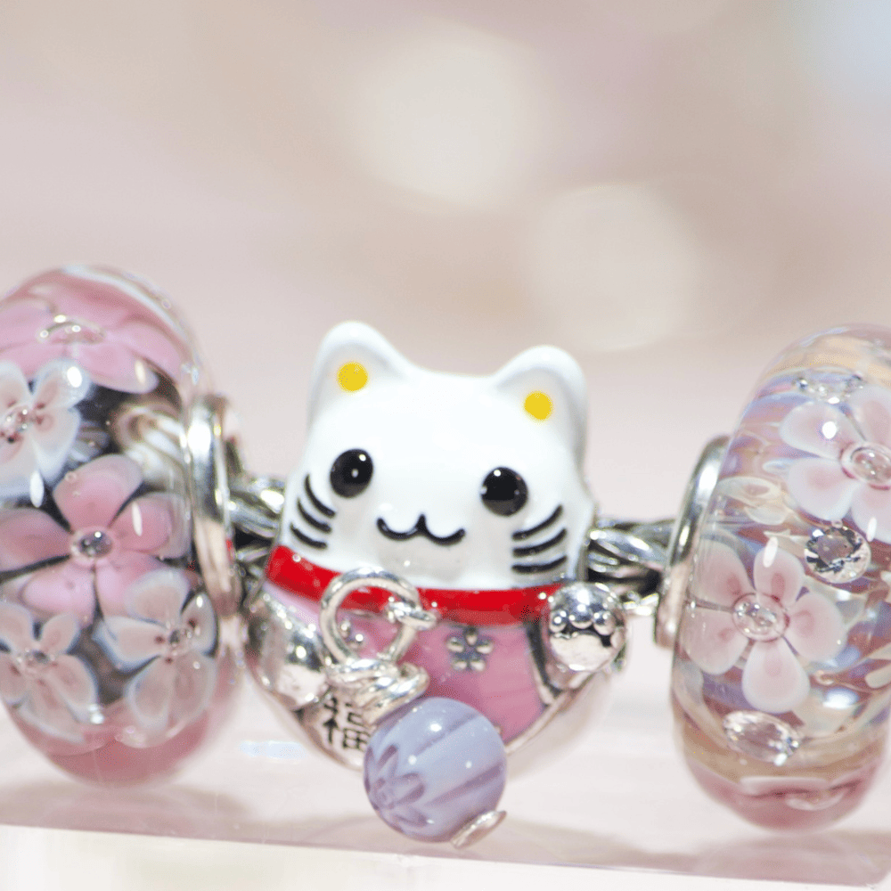 Macro shot of a silver Maneki Neko charm showing a DIY repair: a tiny colorful millefleur glass bead attached with silver wire replacing the original missing bell.
