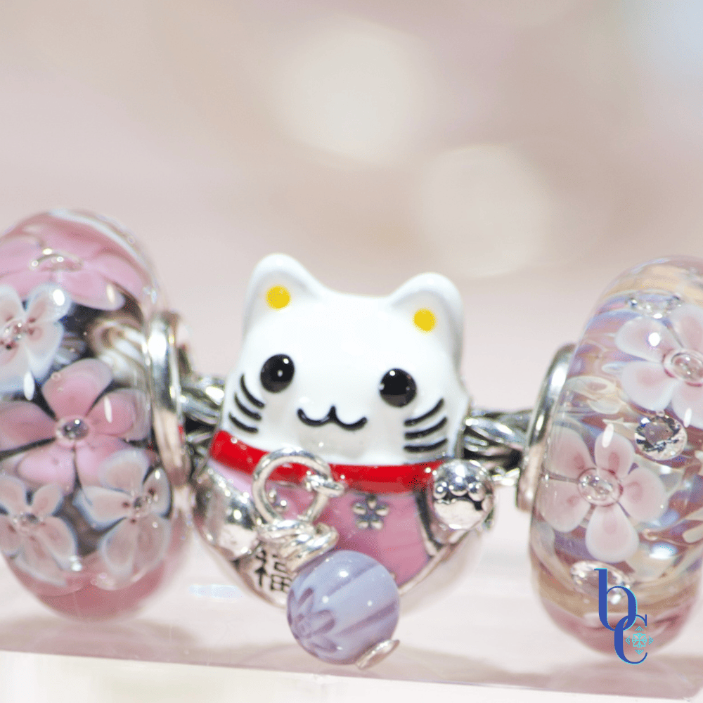 Macro shot of a silver Maneki Neko charm showing a DIY repair: a tiny colorful millefleur glass bead attached with silver wire replacing the original missing bell.