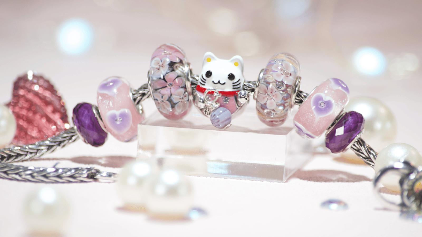 A whimsical European charm bracelet featuring a silver Maneki Neko cat charm, surrounded by pink glass beads, faux pearls, and faux diamonds on a sparkling pink background.