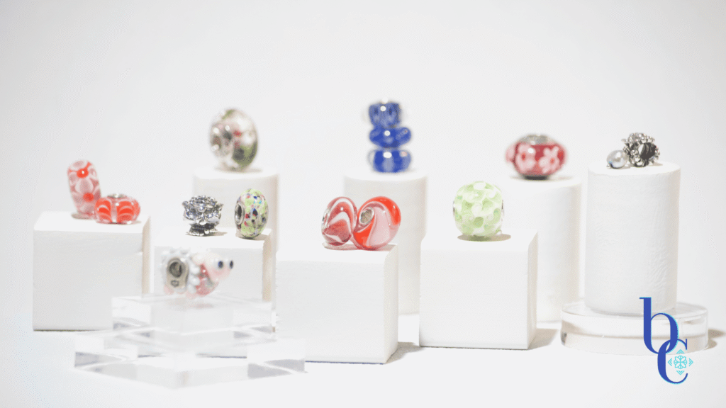 A gallery-style display of rare and OOAK charm beads arranged on clear acrylic risers against a white background, resembling a miniature museum exhibit.