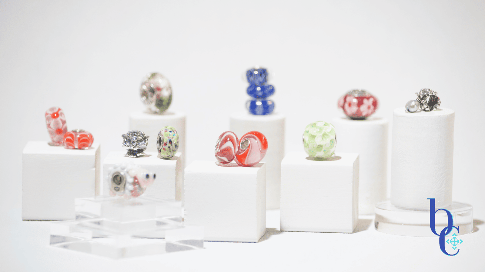 A gallery-style display of rare and OOAK charm beads arranged on clear acrylic risers against a white background, resembling a miniature museum exhibit.