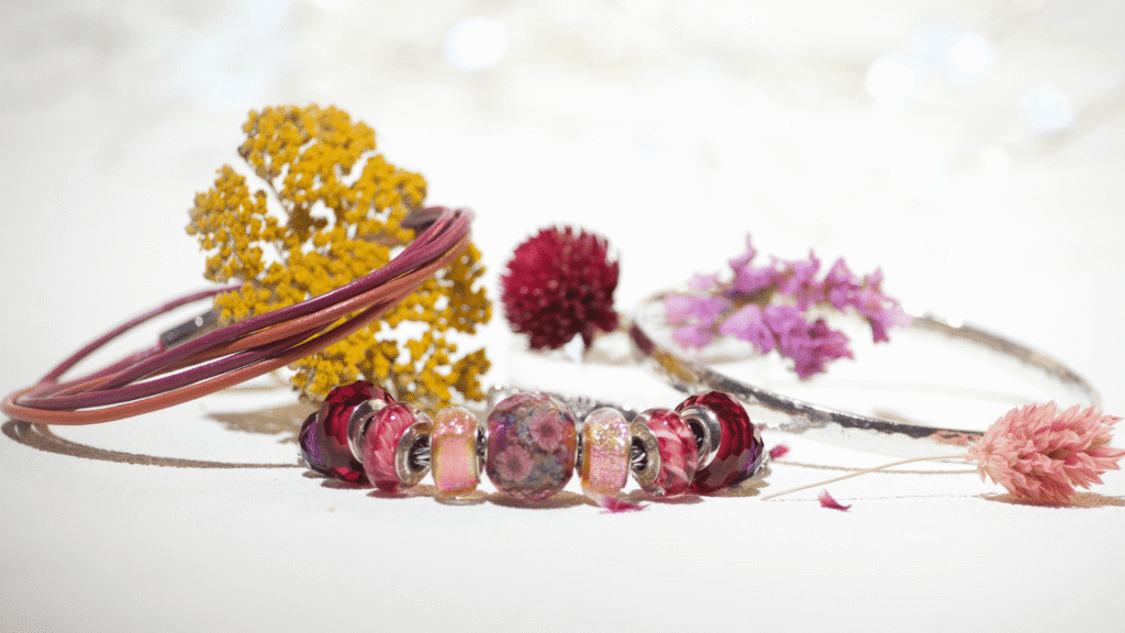 A flat lay photograph showing three separate bracelets laid out neatly on a beige linen surface: a European silver charm bracelet loaded with colorful glass beads in magenta and orange dichroic glass, a magenta and orange thin double wrap leather bracelet with a highly polished hammered silver bangle 