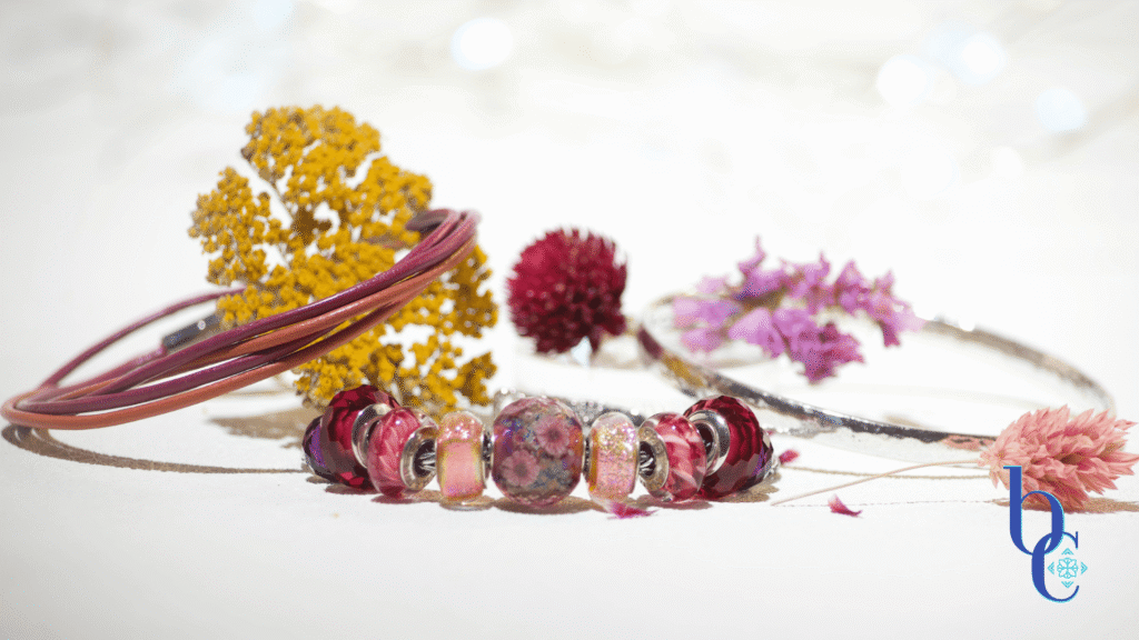 A flat lay photograph showing three separate bracelets laid out neatly on a beige linen surface: a European silver charm bracelet loaded with colorful glass beads in magenta and orange dichroic glass, a magenta and orange thin double wrap leather bracelet with a highly polished hammered silver bangle