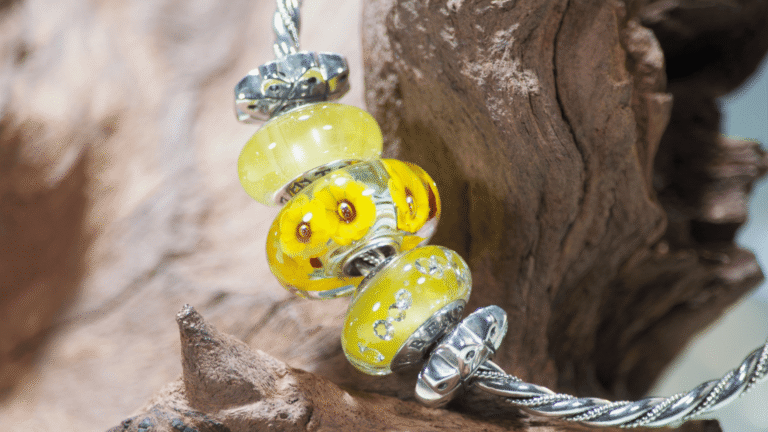 A twisted sterling silver bangle bracelet hanging from a piece of rustic driftwood against a blurred blue background. It features bright yellow and brown sunflower glass Murano beads.