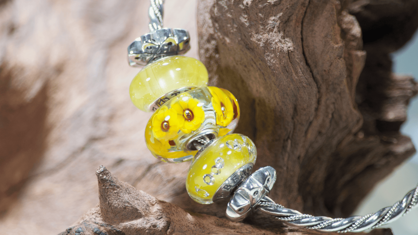 A twisted sterling silver bangle bracelet hanging from a piece of rustic driftwood against a blurred blue background. It features bright yellow and brown sunflower glass Murano beads.