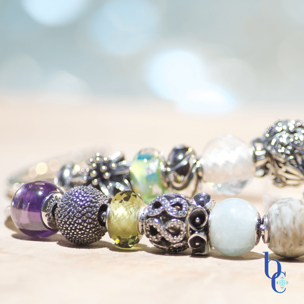 A macro photo of  oxidized silver charms amid assorted colored glass petite European bracelet beads arranged on a rustic pale olivewood slab with a pale blue background.
