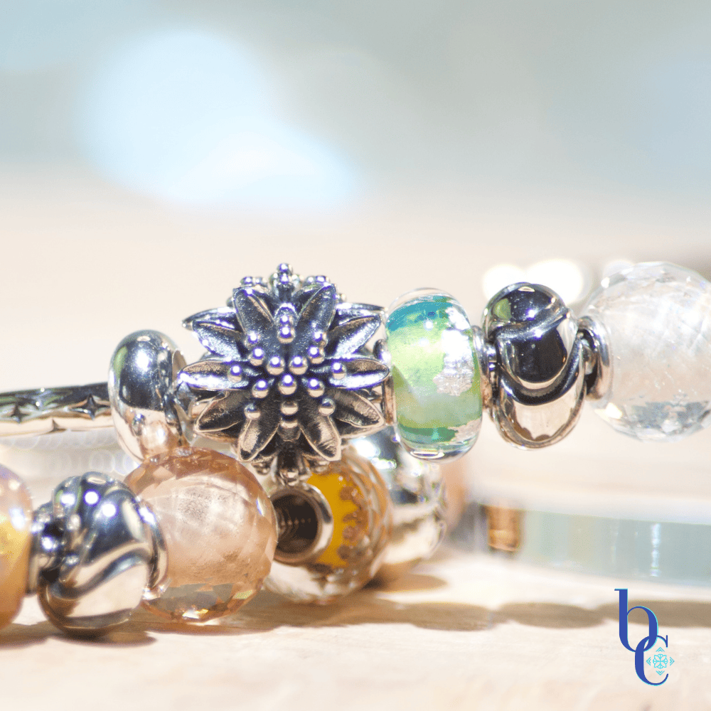 A macro photo of an oxidized silver European flower charm amid assorted colored glass petite European bracelet beads arranged on a rustic pale olivewood slab with a pale blue background.
