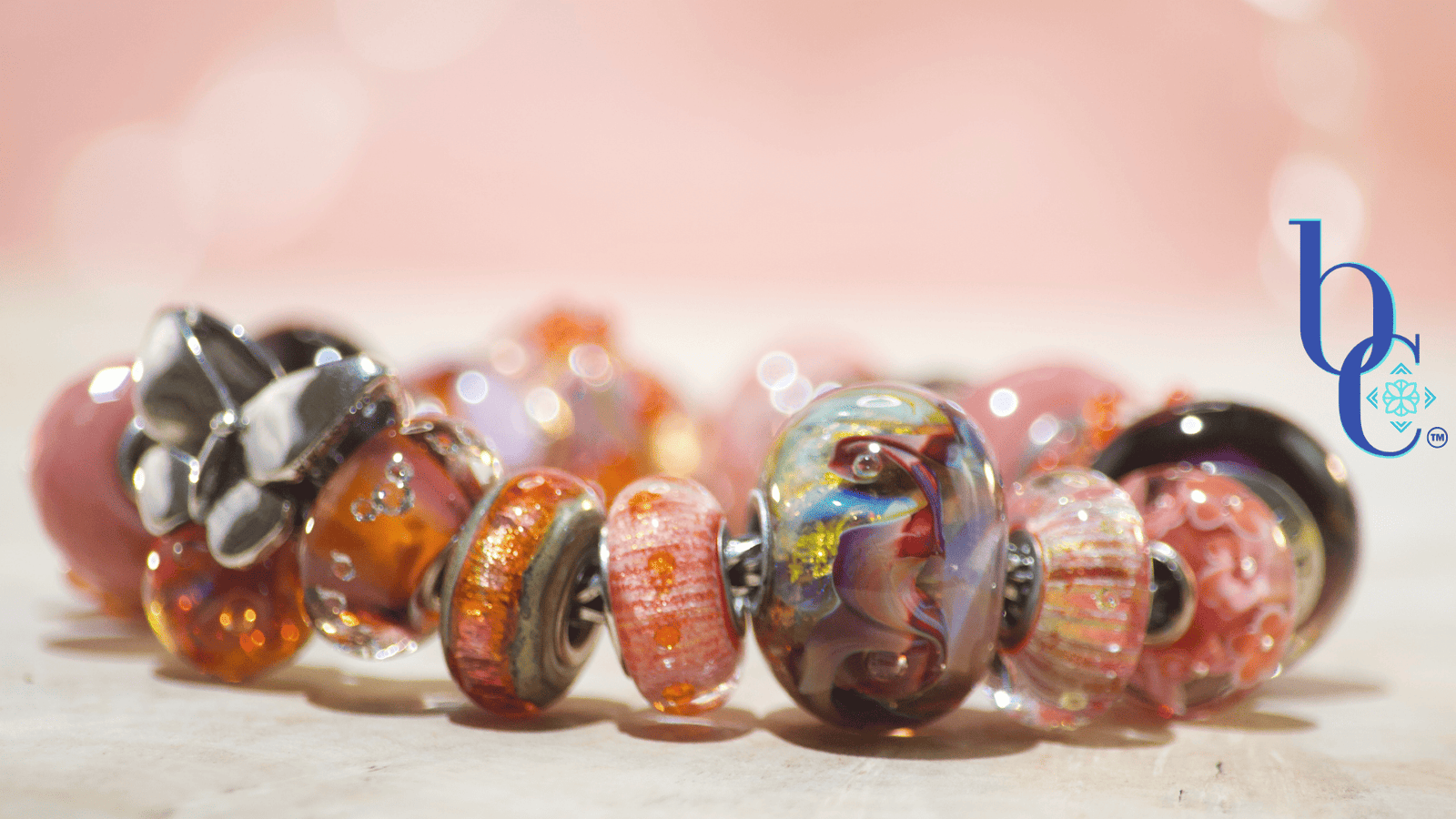 Close-up macro shot of the warm-toned Zuma artisan glass charm bracelet resting on a textured surface. A weighty silver butterfly charm sits asymmetrically among glowing glass beads, including textured orange production beads with internal bubbles, smooth blush pink glass, and a large, swirled cabernet and gold artisan focal. The background is a soft terracotta pink with luminous bokeh.