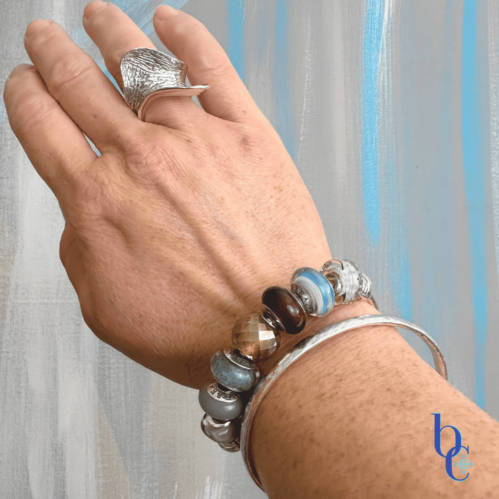 On the Wrist Close-up of a silver charm bangle on olivewood featuring grey, gold, and plum glass beads including TrueBeadz and Ohm designs.