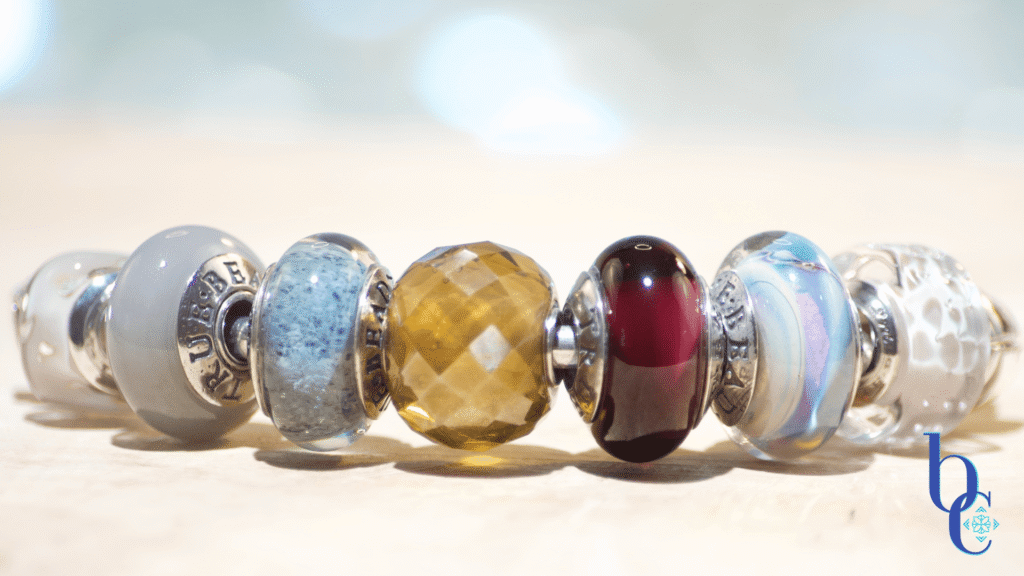 Close-up of a silver charm bangle on olivewood featuring grey, gold, and plum glass beads including TrueBeadz and Ohm designs.