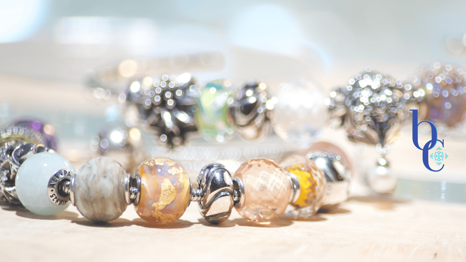 An assortment of petite nano European bracelet beads in glass and silver featuring Elfpetites and a few Pandora Essence pieces arranged on rustic pale olivewood with a blue background.