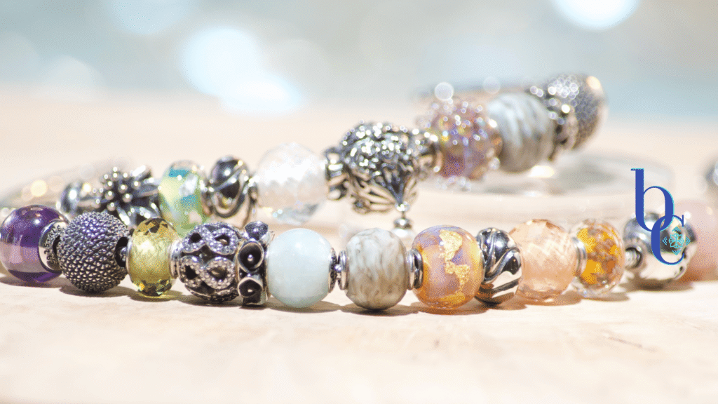 An assortment of petite nano European bracelet beads in glass and silver featuring Elfpetites and a few Pandora Essence pieces arranged on rustic pale olivewood with a blue background.