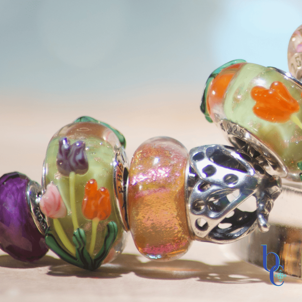 A close-up macro photograph of a silver snake chain charm bracelet resting on tiered, clear acrylic display disks over a textured, raw wood surface. The bracelet features a curated stack of European-style beads, including faceted purple amethyst stones, smooth glass beads with raised orange and purple tulips, a swirling pink and gold glass bead, and an oxidized silver floral spacer. The background is a soft, out-of-focus teal and water-blue bokeh.