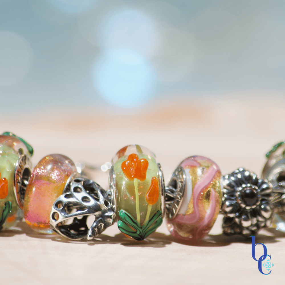 A close-up macro photograph of a silver snake chain charm bracelet resting on tiered, clear acrylic display disks over a textured, raw wood surface. The bracelet features a curated stack of European-style beads, including faceted purple amethyst stones, smooth glass beads with raised orange and purple tulips, a swirling pink and gold glass bead, and an oxidized silver floral spacer. The background is a soft, out-of-focus teal and water-blue bokeh.