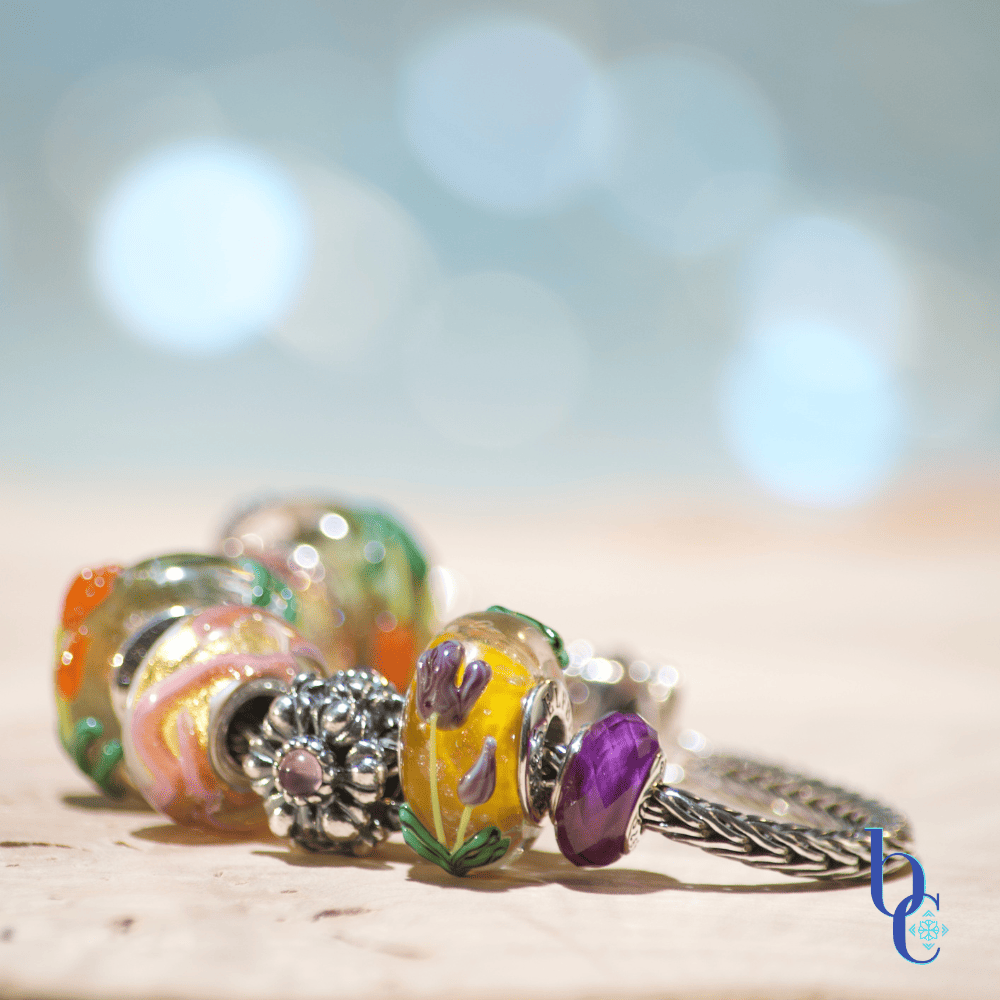 A close-up macro photograph of a silver snake chain charm bracelet resting on tiered, clear acrylic display disks over a textured, raw wood surface. The bracelet features a curated stack of European-style beads, including faceted purple amethyst stones, smooth glass beads with raised orange and purple tulips, a swirling pink and gold glass bead, and an oxidized silver floral spacer. The background is a soft, out-of-focus teal and water-blue bokeh.