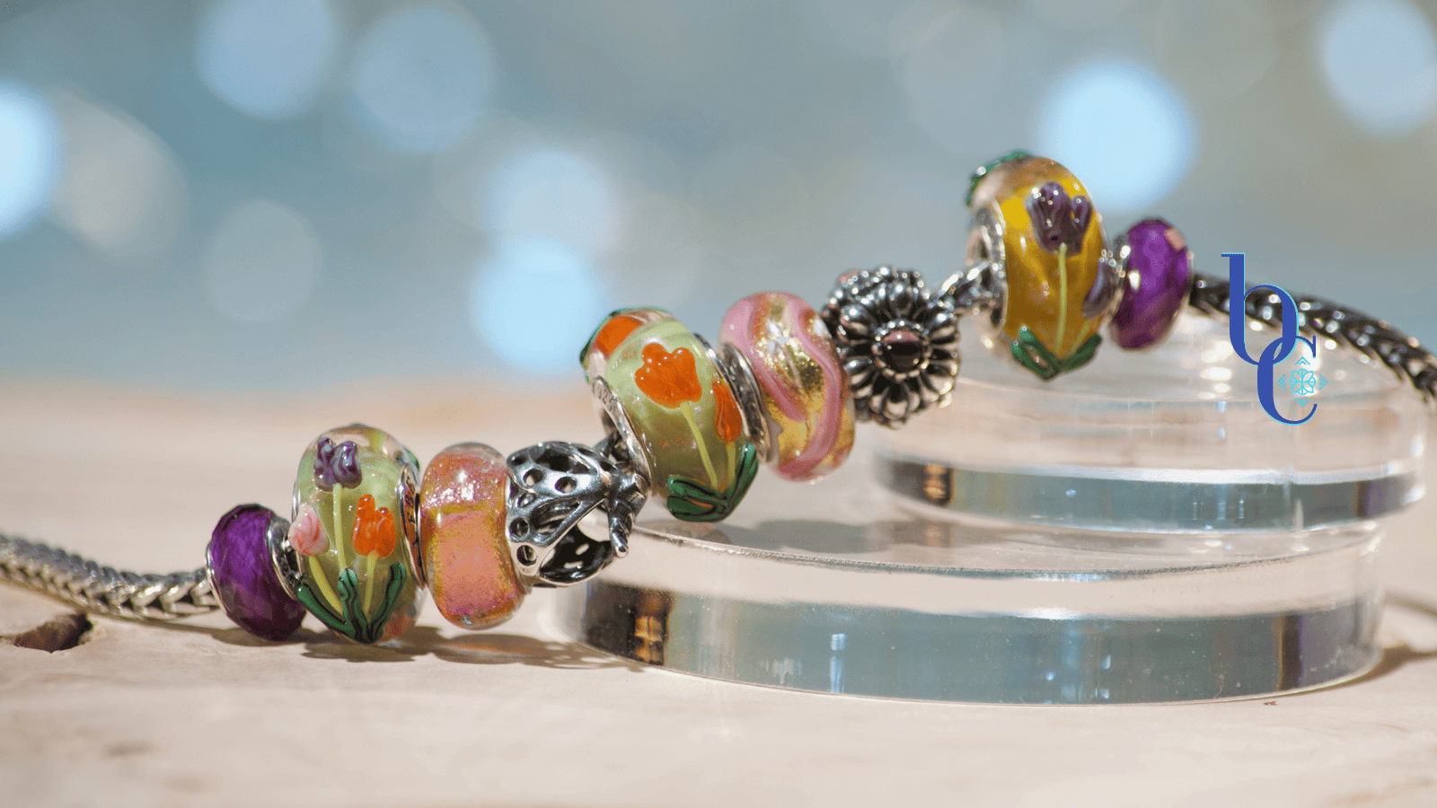 A close-up macro photograph of a silver snake chain charm bracelet resting on tiered, clear acrylic display disks over a textured, raw wood surface. The bracelet features a curated stack of European-style beads, including faceted purple amethyst stones, smooth glass beads with raised orange and purple tulips, a swirling pink and gold glass bead, and an oxidized silver floral spacer. The background is a soft, out-of-focus teal and water-blue bokeh.