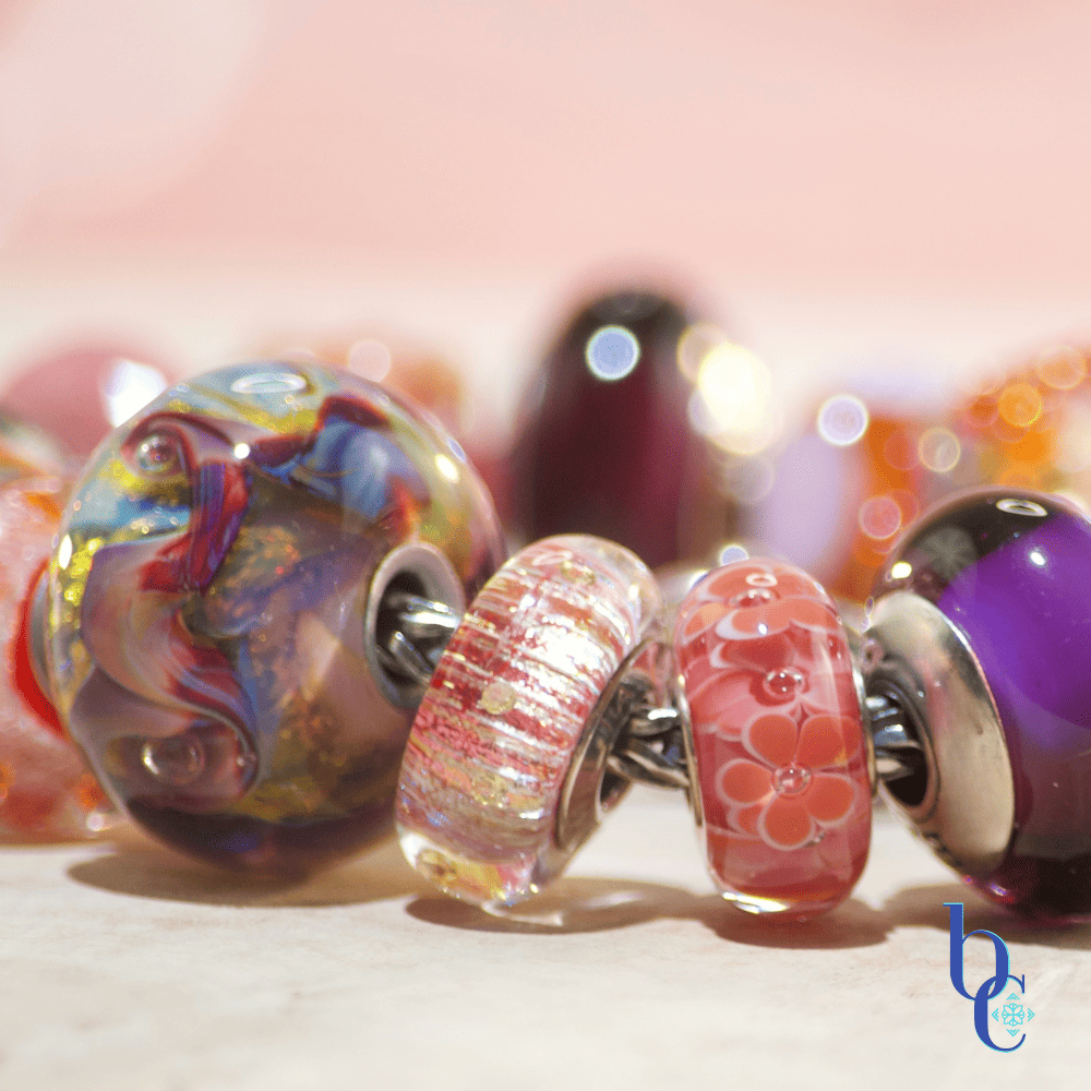 Close-up macro shot of the warm-toned Zuma artisan glass charm bracelet resting on a textured surface. A weighty silver butterfly charm sits asymmetrically among glowing glass beads, including textured orange production beads with internal bubbles, smooth blush pink glass, and a large, swirled cabernet and gold artisan focal. The background is a soft terracotta pink with luminous bokeh.
