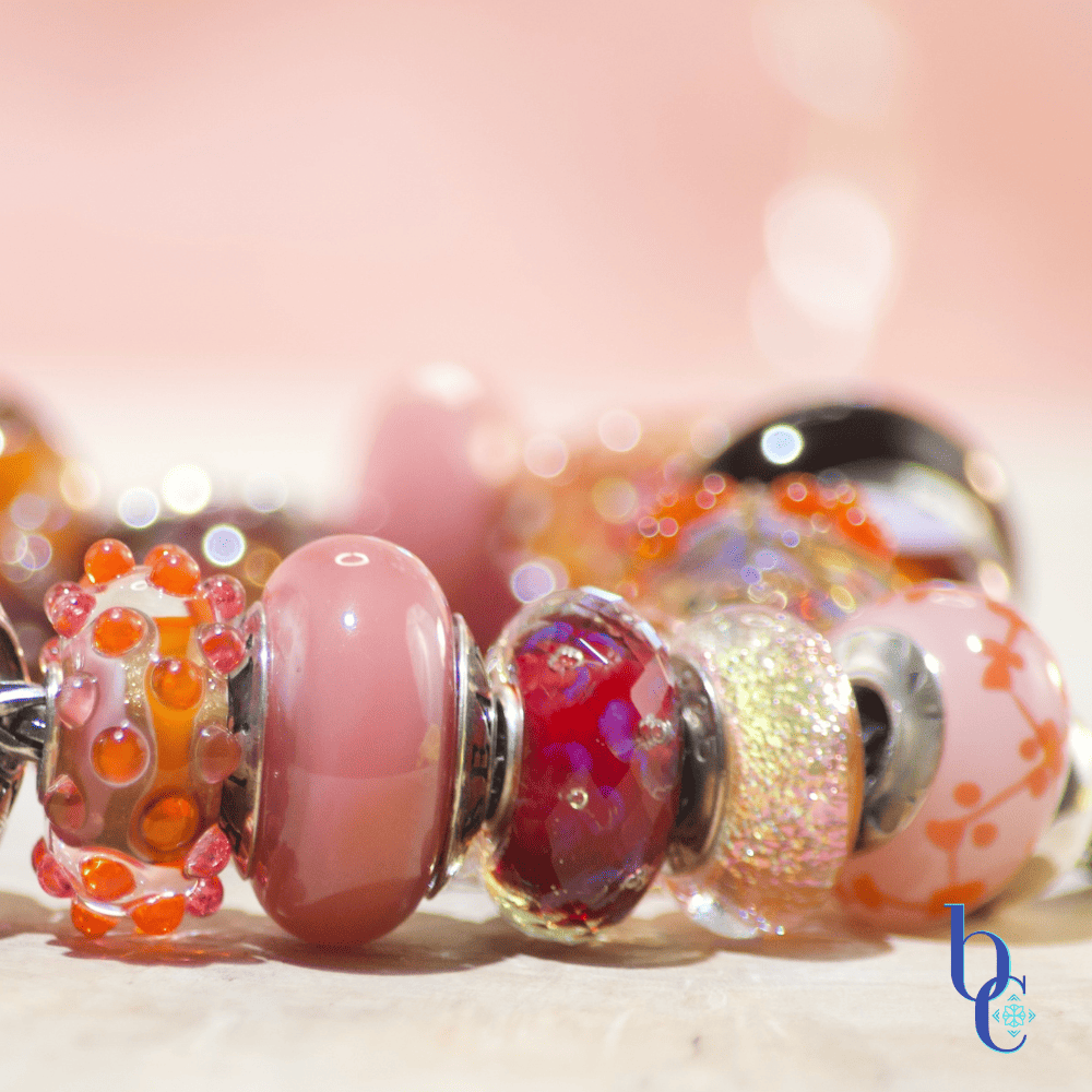 Close-up macro shot of the warm-toned Zuma artisan glass charm bracelet resting on a textured surface. A weighty silver butterfly charm sits asymmetrically among glowing glass beads, including textured orange production beads with internal bubbles, smooth blush pink glass, and a large, swirled cabernet and gold artisan focal. The background is a soft terracotta pink with luminous bokeh.