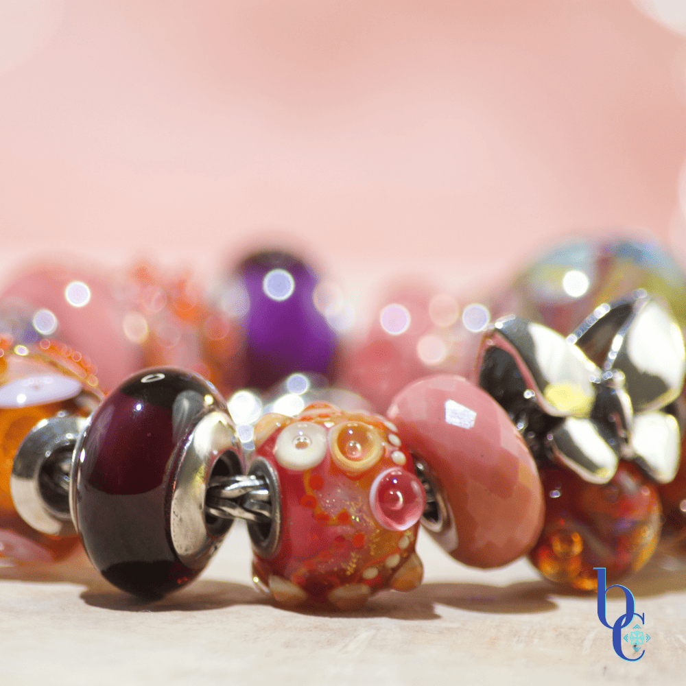 Close-up macro shot of the warm-toned Zuma artisan glass charm bracelet resting on a textured surface. A weighty silver butterfly charm sits asymmetrically among glowing glass beads, including textured orange production beads with internal bubbles, smooth blush pink glass, and a large, swirled cabernet and gold artisan focal. The background is a soft terracotta pink with luminous bokeh.