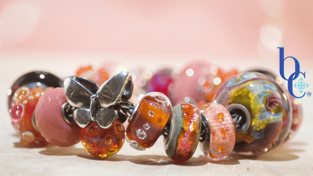 Close-up macro shot of the warm-toned Zuma artisan glass charm bracelet resting on a textured surface. A weighty silver butterfly charm sits asymmetrically among glowing glass beads, including textured orange production beads with internal bubbles, smooth blush pink glass, and a large, swirled cabernet and gold artisan focal. The background is a soft terracotta pink with luminous bokeh.