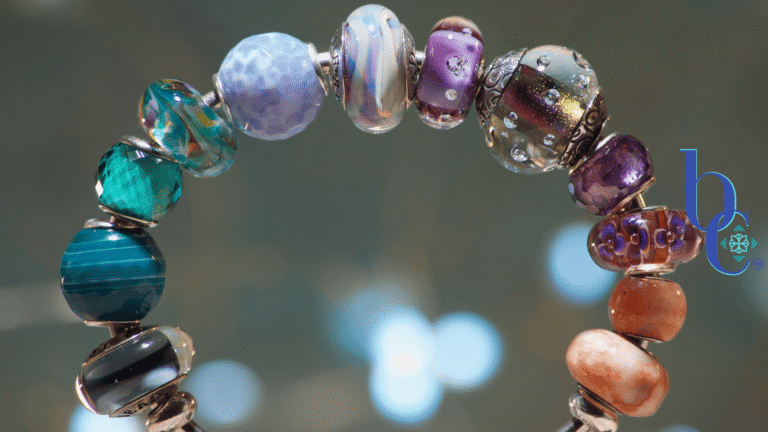 A macro image of a silver bangle bracelet filled with glass beads in tones from blue and aqua through amethyst and tangerine set against a moody grey backdrop with pale bokeh accents.