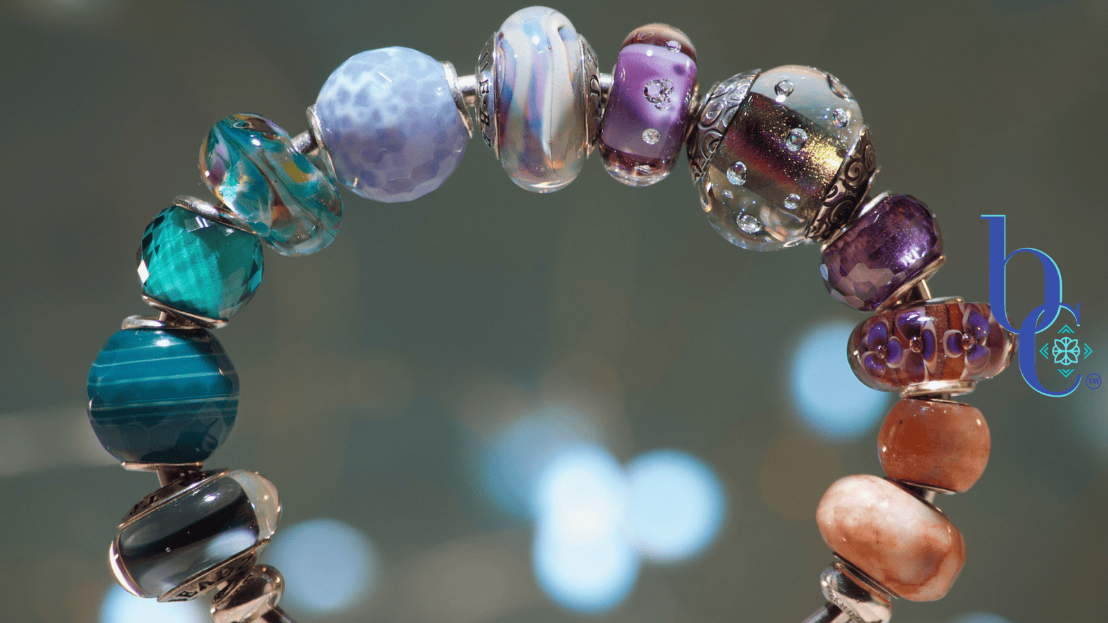 A macro image of a silver bangle bracelet filled with glass beads in tones from blue and aqua through amethyst and tangerine set against a moody grey backdrop with pale bokeh accents.