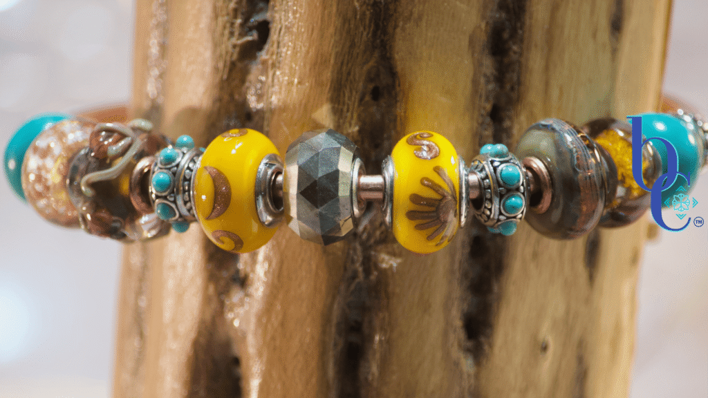 Close-up macro shot of a copper charm bangle resting against the deeply textured, porous surface of native desert cholla wood. At the center is a large, faceted natural golden pyrite bead, flanked by bright sunflower-yellow Black Raven Beads featuring coppery sun and moon motifs. The design radiates outward with deeply oxidized silver spacers dotted with bright turquoise, followed by brown floral lampwork glass with copper sparkle, and finishes with smooth, bright turquoise end beads.