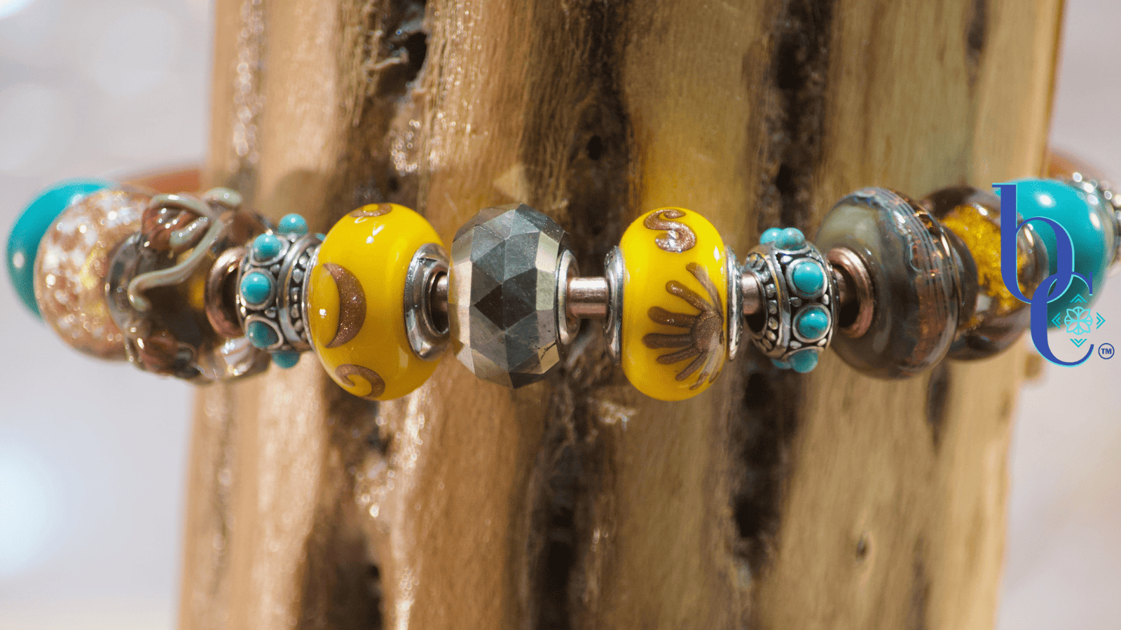 Close-up macro shot of a copper charm bangle resting against the deeply textured, porous surface of native desert cholla wood. At the center is a large, faceted natural golden pyrite bead, flanked by bright sunflower-yellow Black Raven Beads featuring coppery sun and moon motifs. The design radiates outward with deeply oxidized silver spacers dotted with bright turquoise, followed by brown floral lampwork glass with copper sparkle, and finishes with smooth, bright turquoise end beads.