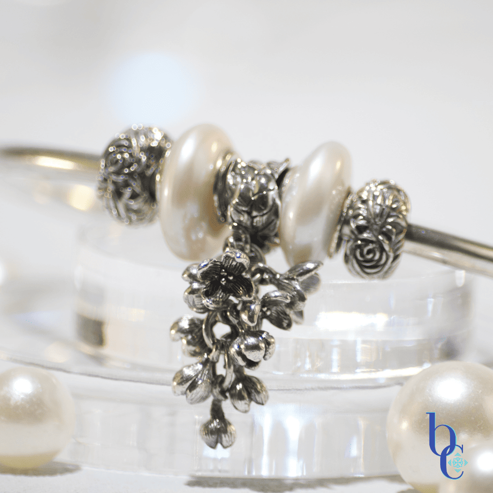 Close-up of a minimalist silver charm bangle resting on clear acrylic tiers alongside scattered white pearls. The central focal is an antique oxidized silver OHM Beads 'Lilac Dreams' floral dangle, flanked by two irregular baroque coin pearls and silver rose stoppers. The background is a soft, misty grey bathed in an atmospheric amethyst purple light that subtly reflects on the edges of the pearls. The Bijoux Chat logo is in the right corner.