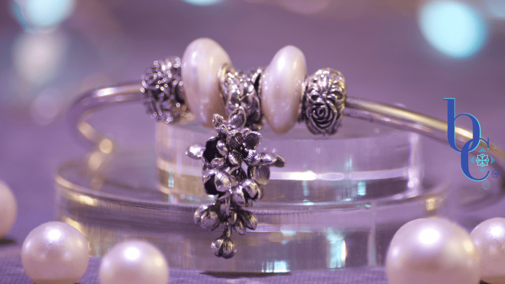Close-up of a minimalist silver charm bangle resting on clear acrylic tiers alongside scattered white pearls. The central focal is an antique oxidized silver OHM Beads 'Lilac Dreams' floral dangle, flanked by two irregular baroque coin pearls and silver rose stoppers. The background is a soft, misty grey bathed in an atmospheric amethyst purple light that subtly reflects on the edges of the pearls. The Bijoux Chat logo is in the right corner.
