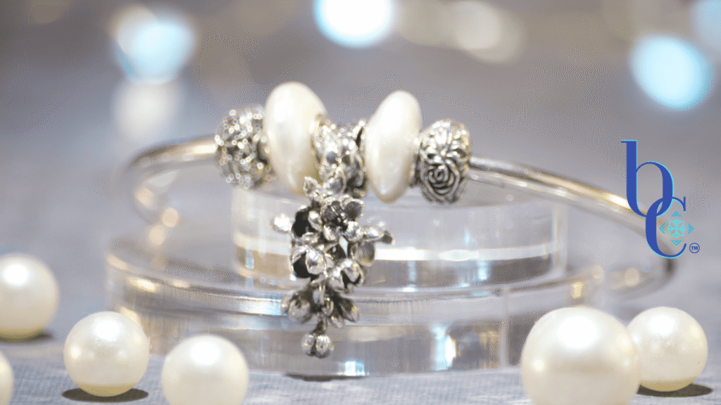Close-up of a minimalist silver charm bangle resting on clear acrylic tiers alongside scattered white pearls. The central focal is an antique oxidized silver OHM Beads 'Lilac Dreams' floral dangle, flanked by two irregular baroque coin pearls and silver rose stoppers. The background is a soft, misty grey bathed in an atmospheric amethyst purple light that subtly reflects on the edges of the pearls. The Bijoux Chat logo is in the right corner.