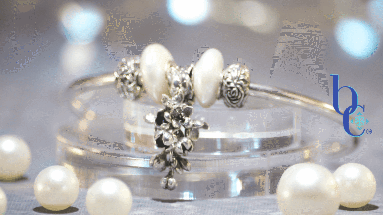 Close-up of a minimalist silver charm bangle resting on clear acrylic tiers alongside scattered white pearls. The central focal is an antique oxidized silver OHM Beads 'Lilac Dreams' floral dangle, flanked by two irregular baroque coin pearls and silver rose stoppers. The background is a soft, misty grey bathed in an atmospheric amethyst purple light that subtly reflects on the edges of the pearls. The Bijoux Chat logo is in the right corner.
