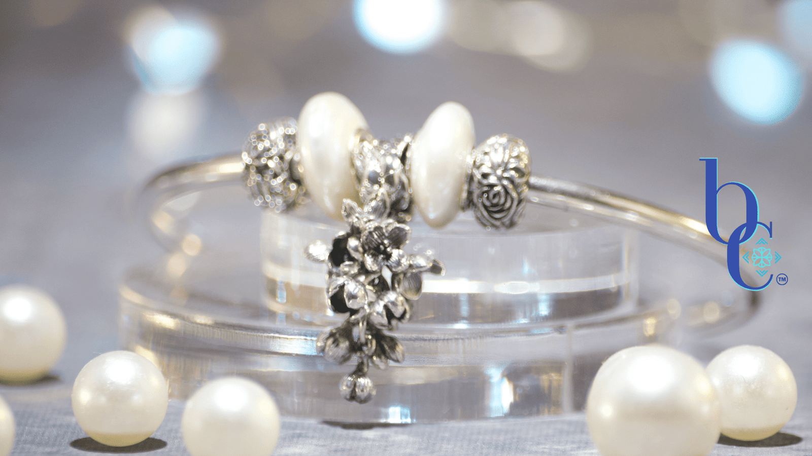 Close-up of a minimalist silver charm bangle resting on clear acrylic tiers alongside scattered white pearls. The central focal is an antique oxidized silver OHM Beads 'Lilac Dreams' floral dangle, flanked by two irregular baroque coin pearls and silver rose stoppers. The background is a soft, misty grey bathed in an atmospheric amethyst purple light that subtly reflects on the edges of the pearls. The Bijoux Chat logo is in the right corner.