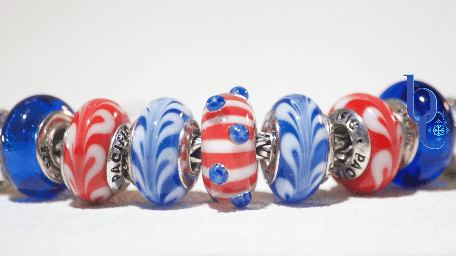 A minimalist flat lay of the Trollbeads Star Spangled bead flanked by deep red and true blue glass charms, resting on a stark, crisp white background.